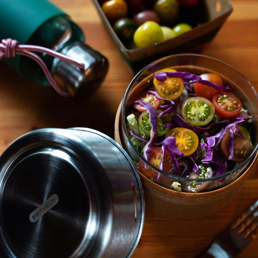 Runde Glas Bowl mit Tomatensalat befüllt auf einem Tisch stehend neben Edelstahl Deckel und liegender Trinkflasche