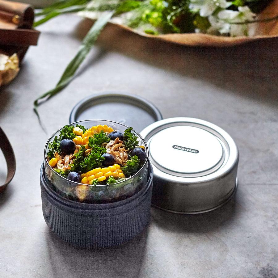 Runde Lunch Bowl aus Glas auf Tisch stehend mit schieferfarbener Schutzhülle und Salat befüllt