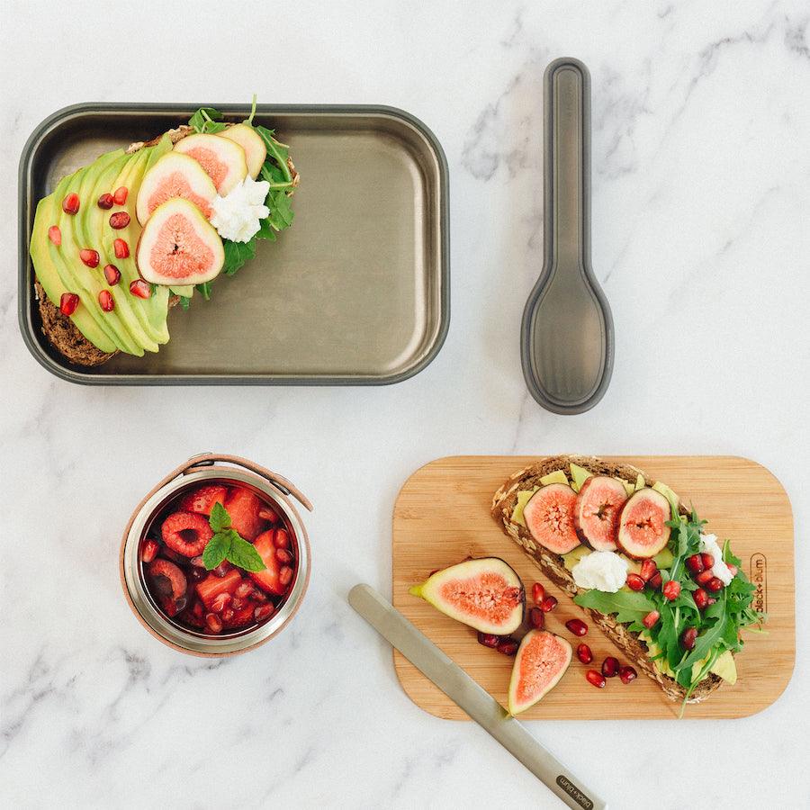 Geöffnete Sandwich Box aus Edelstahl mit Brot und Avocado und danebenliegendem Bambusdeckel mit ebenfalls Brot darauf