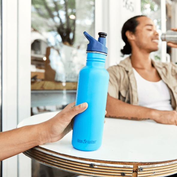 Person hält blaue Edelstahl Trinkflasche in der Hand auf dem Tisch mit trinkendem Mann im Hintergrund