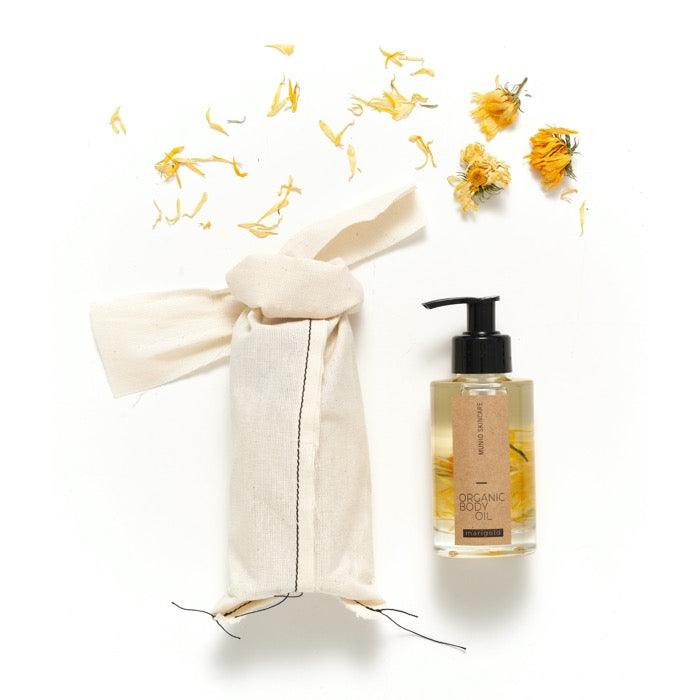 Bio Body Öl in Flasche mit Spender neben Stoffverpackung und Ringelblumenblüten
