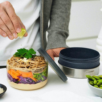 Person drückt Limette über runde Bowl aus Glas aus, die mit Nudeln und Gemüse befüllt ist