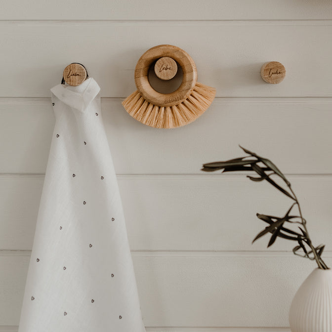 Drei Knäufe aus Holz an einer weißen Wand hängend mit einem weißem Küchentuch und einer Gemüsebürste daran hängend