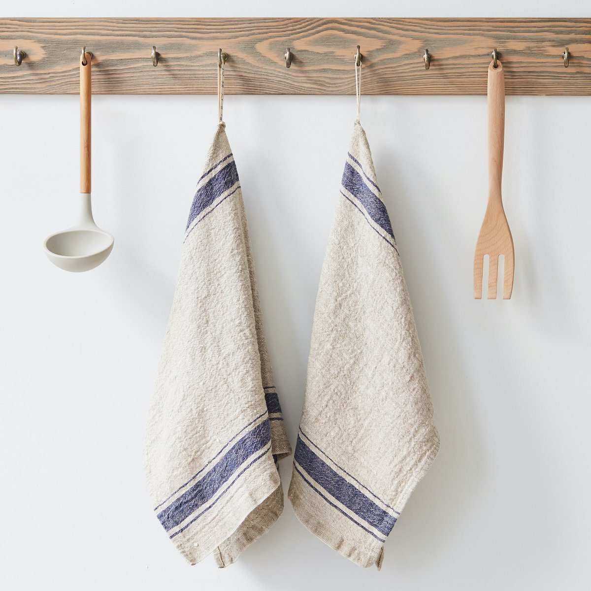 Zwei Küchentücher aus Leinen mit blauen Streifen hängen neben einer Kelle und einer Gabel an einem Holzbrett